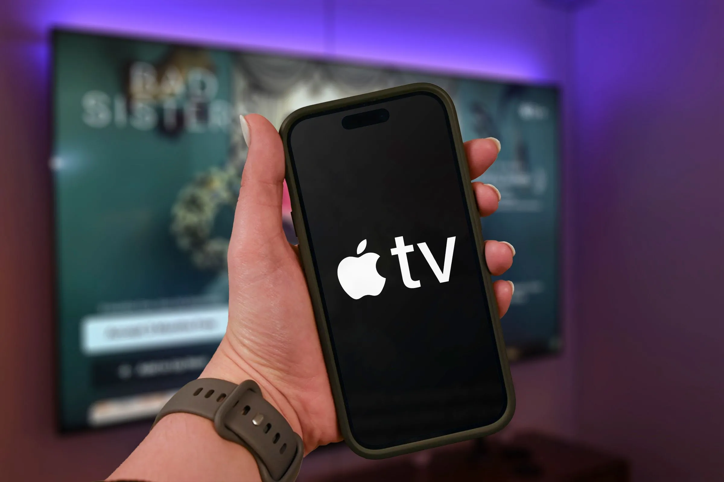 Bàn tay cầm iPhone hiển thị logo Apple TV+, phía sau là TV đang phát nội dung.