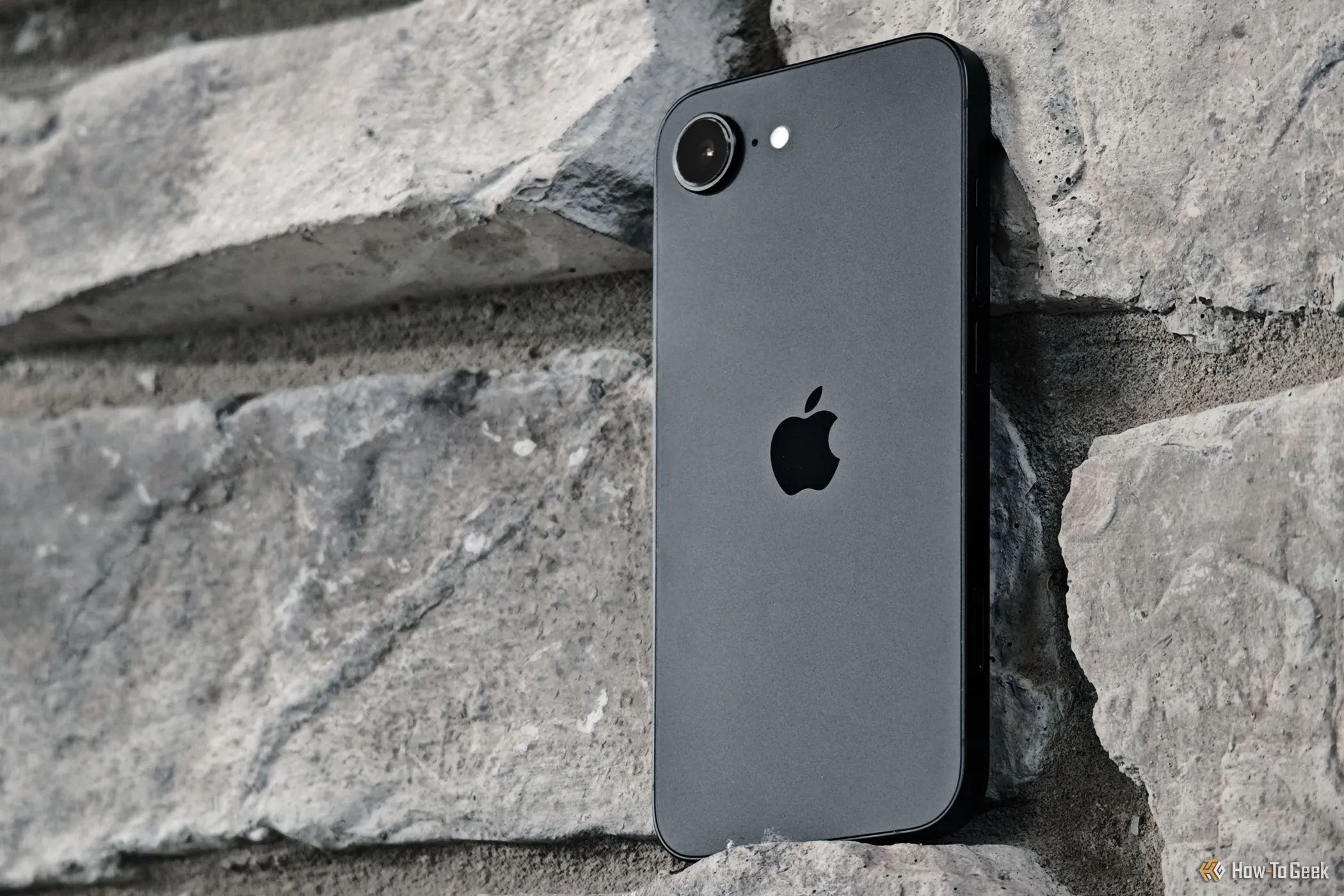 Chiếc điện thoại iPhone 16e được đặt dựa vào bức tường gạch, làm nổi bật thiết kế và hệ thống camera đơn của thiết bị
