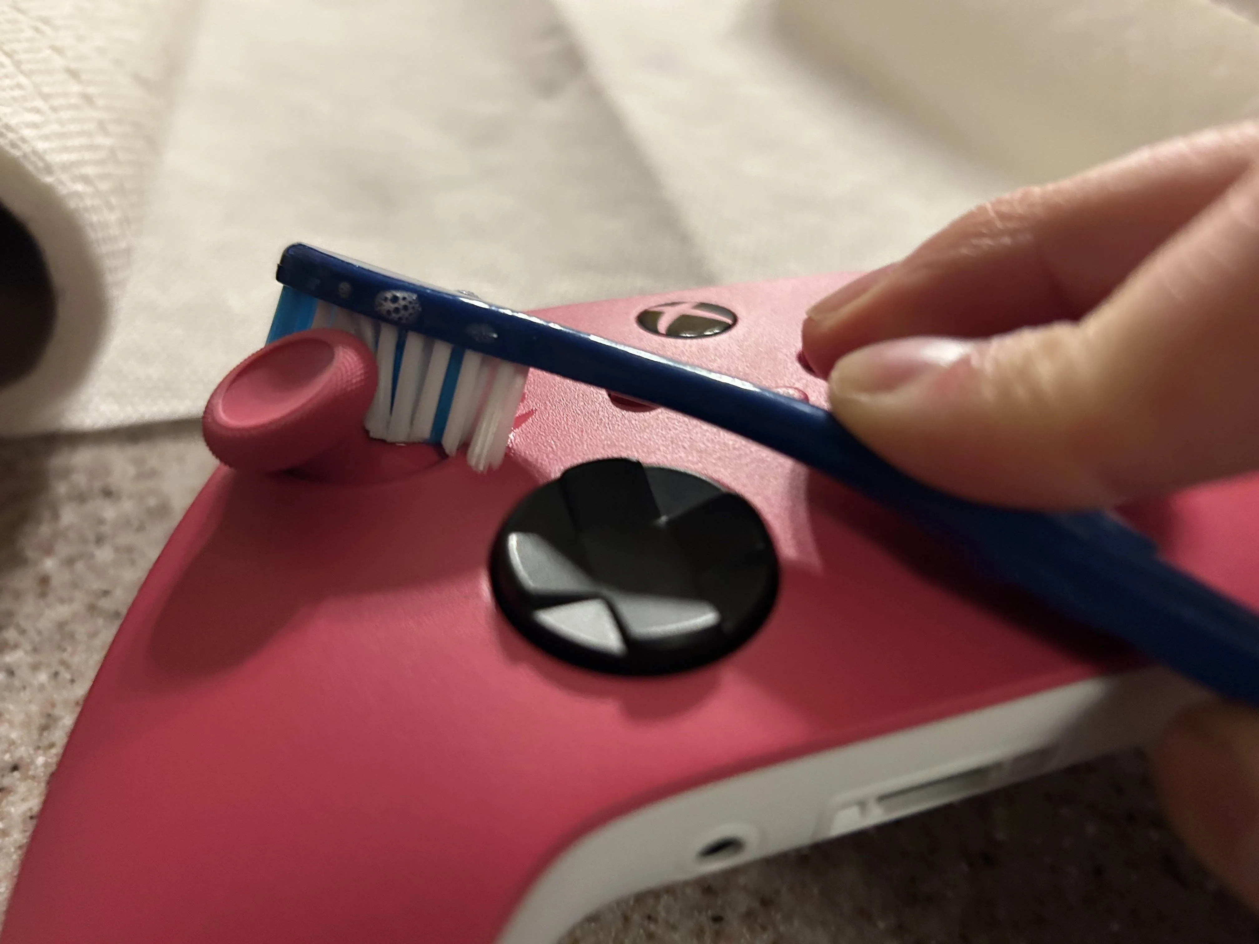 Dùng bàn chải màu xanh làm sạch các cạnh của cần analog trên tay cầm Xbox màu hồng để loại bỏ bụi bẩn.