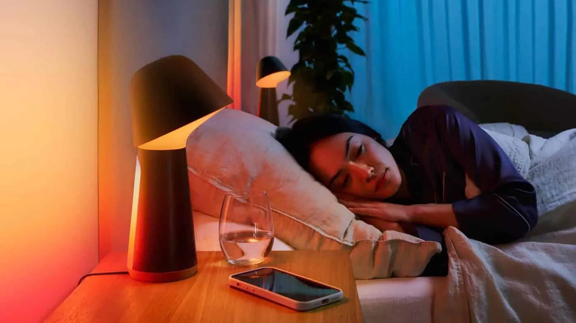 Người phụ nữ đang ngủ trên giường cạnh đèn thông minh và điện thoại smartphone.