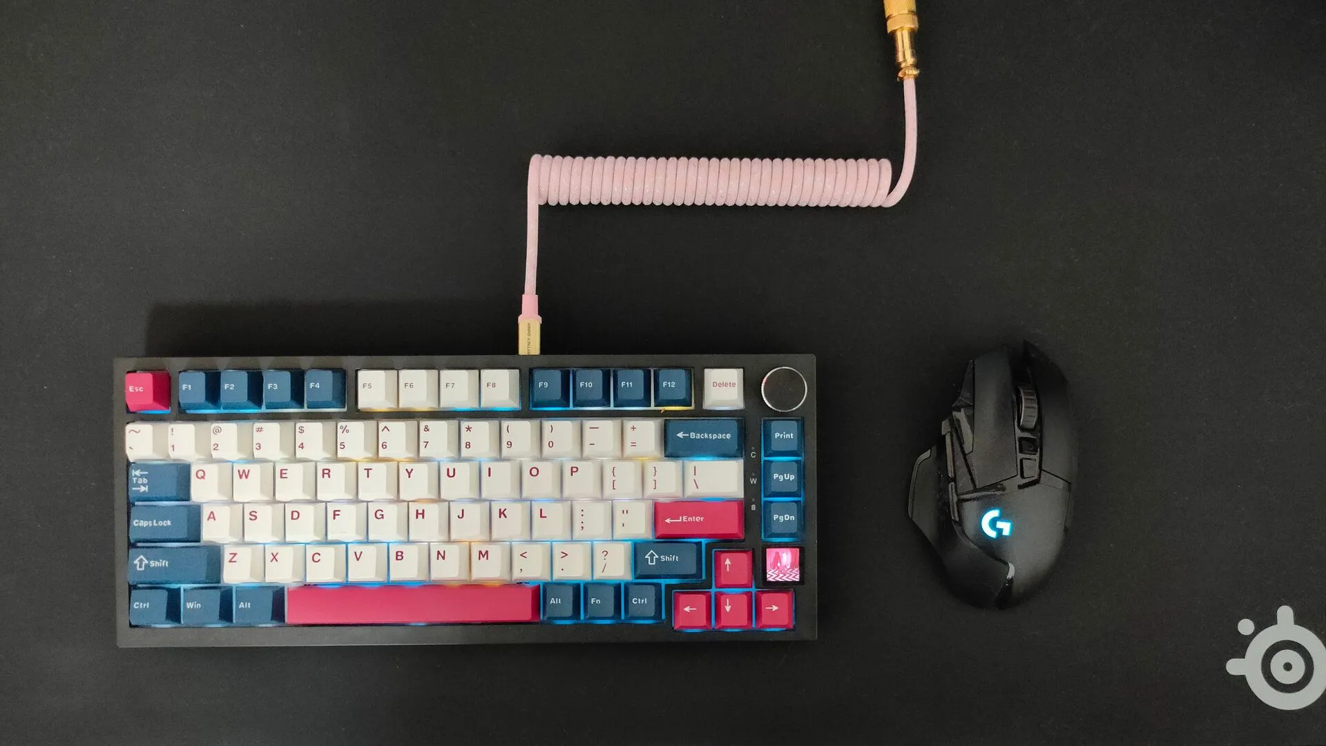 Bàn phím cơ có dây với cáp xoắn màu hồng nổi bật bên cạnh chuột không dây Logitech G502, thể hiện sự kết hợp thiết bị có dây và không dây trên bàn làm việc.