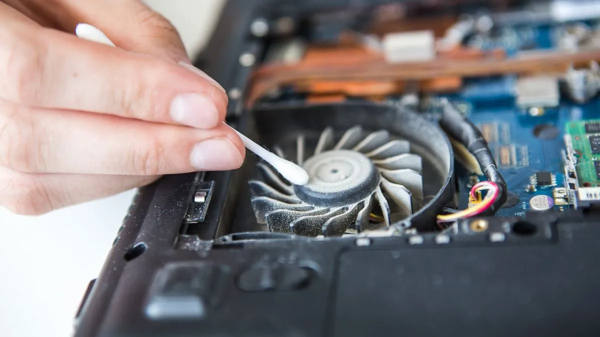 Một người đang dùng tăm bông để làm sạch bụi từ khe tản nhiệt của laptop, minh họa việc vệ sinh thiết bị.