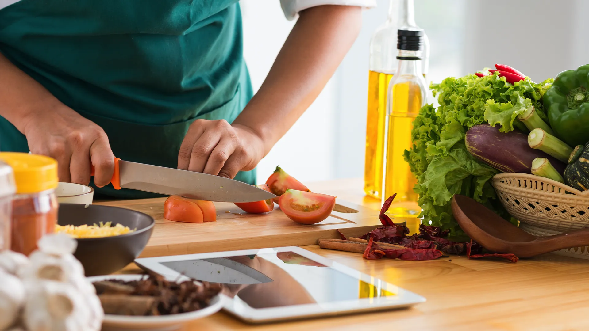 Hình ảnh cận cảnh một người phụ nữ đang thái rau củ trong bếp, trên bàn có một chiếc máy tính bảng hiển thị công thức nấu ăn.