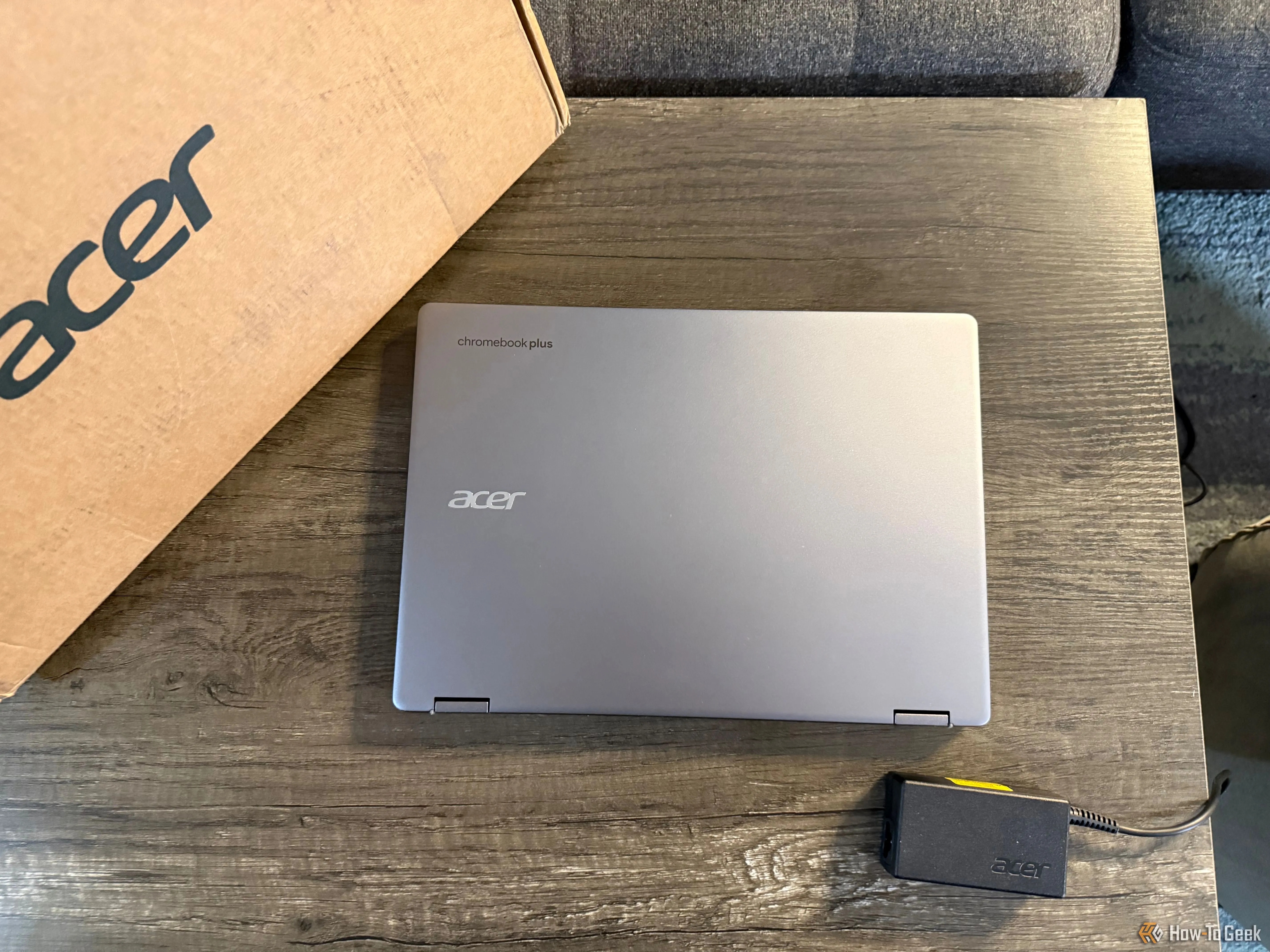 Laptop Acer Chromebook Plus Spin 514 màu bạc đang gập lại, đặt cạnh hộp sản phẩm và bộ sạc trên bề mặt gỗ